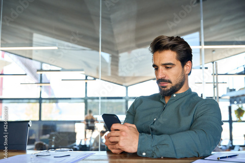 Busy young Latin business man using mobile cell phone at work in office. Serious male executive businessman manager sitting at desk holding smartphone working on cellphone at workplace.