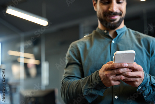 Young business man, businessman employee standing in office holding smartphone in hands using apps on cell phone doing digital payments on cellphone at work. Mobile technology concept. Close up view