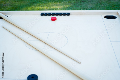 Novuss table with discs and cue stics. Novuss (also known as koroona or korona) is a large wooden board game where small wooden discs are hit with cue sticks into pockets.