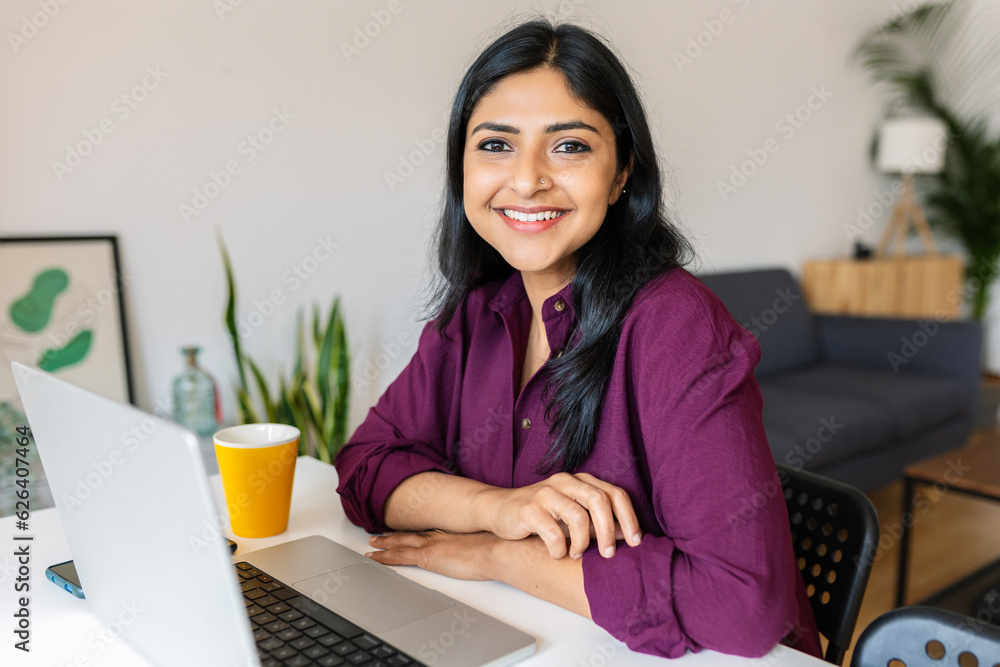 Young indian woman smile at camera working with laptop at home. Confident ethnic female sitting on living room table using computer. - obrazy, fototapety, plakaty 