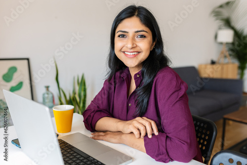 Young indian woman smile at camera working with laptop at home. Confident ethnic female sitting on living room table using computer. photo