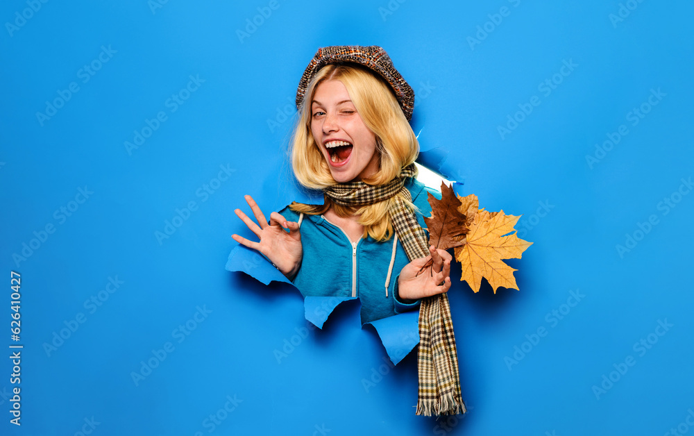 Winking autumn girl in cap and scarf showing ok sign. Happy beautiful woman with autumn leaves looking through paper hole. Autumn sales. Discount. Autumn clothing collection. Fashion trends for fall.