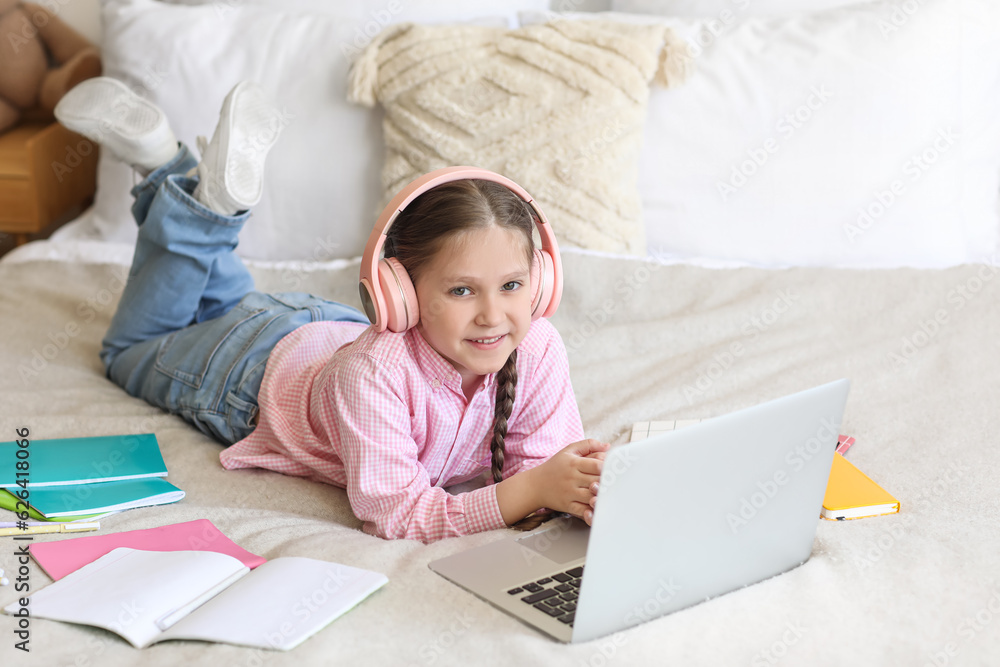 Little girl in headphones with laptop studying computer sciences online on bed at home Little girl in headphones with laptop studying computer sciences online on bed at home