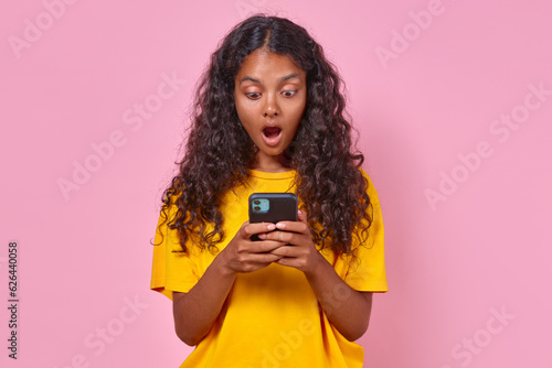 Young surprised Indian woman teenager with phone in hands is shocked by what saw and opens mouth wide reading news about increase in taxes stands posing on pink background. Wow delight