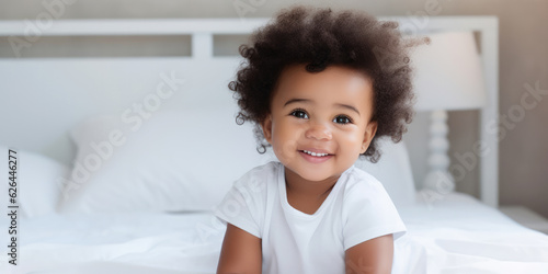 Happy Black Little Baby Girl Sitting On Bed Looking At Camera And Smiling In Bedroom At Home. Advertisement Banner With Cute Child Posing Indoor. Panorama With Copy Space For Your Text