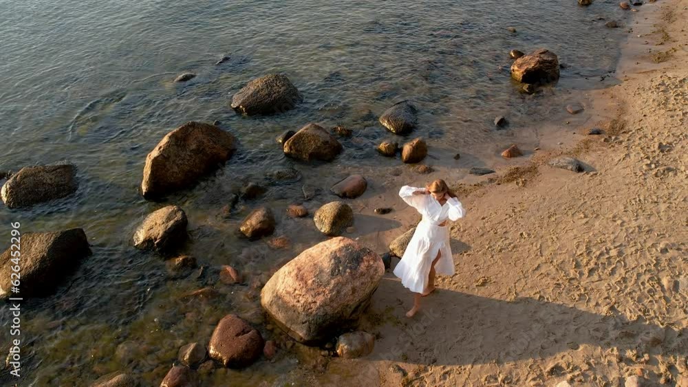 A woman walks along the seashore during sunrise dressed in a flowing white dress - view from above from a drone. A woman walks along the seashore during sunrise dressed in a flowing white dress - view from above from a drone.