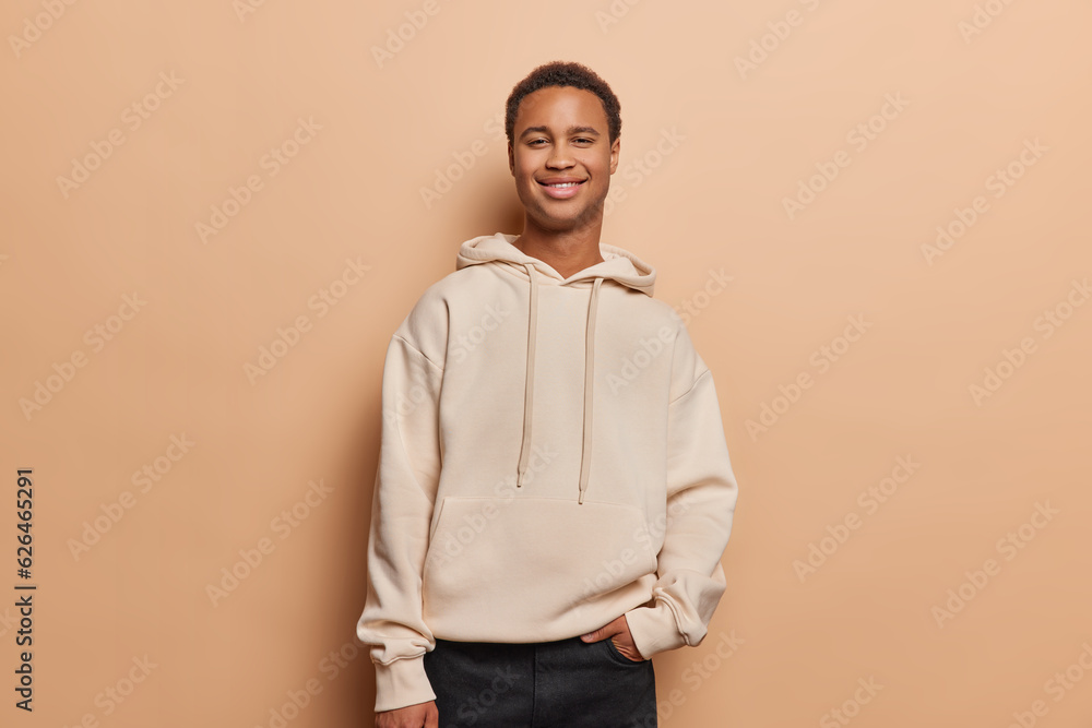 People and happiness. Indoor photo of young cheerful smiling African man standing in centre isolated on beige background looking straight at camera wearing casual clothes keeping hand in pocket