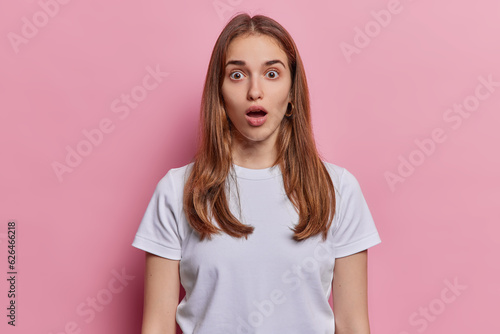 People emotions. Studio waist up of surprised young European girl wearing tshirt shocked seeing something unusual or unexpected keeping hands down standing in centre isolated on pink background