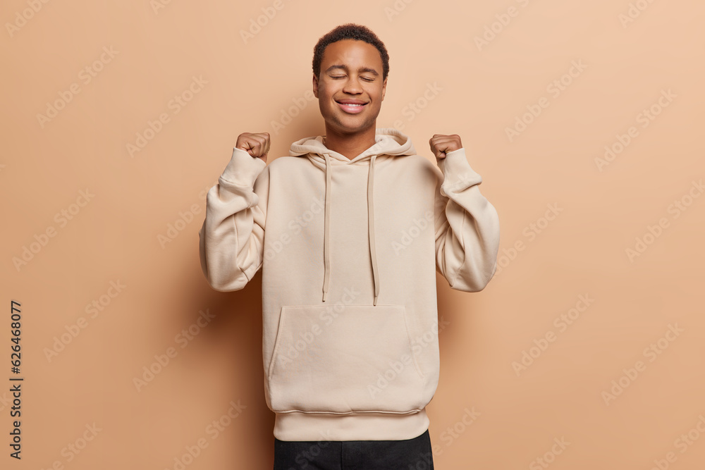 People and positive emotions concept. Studio shot of young cheerful smiling African man with closed eyes poses in centre isolated on beige background wearing casual clothes keeping hands raised People and positive emotions concept. Studio shot of young cheerful smiling African man with closed eyes poses in centre isolated on beige background wearing casual clothes keeping hands raised