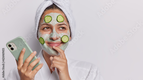 Dreamy young woman holds mobile phone buys beauty products online focused aside with glad expression applies clay facial mask and cucumber slices wears bathrobe and towel isolated on white wall