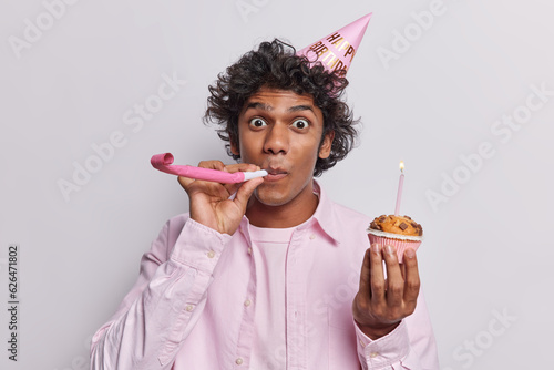 Horizontal shot of handsome curly haired Hindu man blows in party horn holds delicious cupcake with candle wears formal pink shirt celebrates birthday isolated over white background. Holiday concept photo