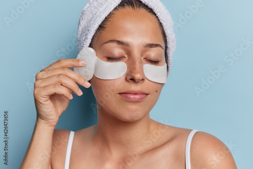 Close up shot of young woman with healthy glowing skin applies beauty patches under eyes and uses face massager undergoes cosmetology procedures wears bath towel on head isolated on blue wall photo