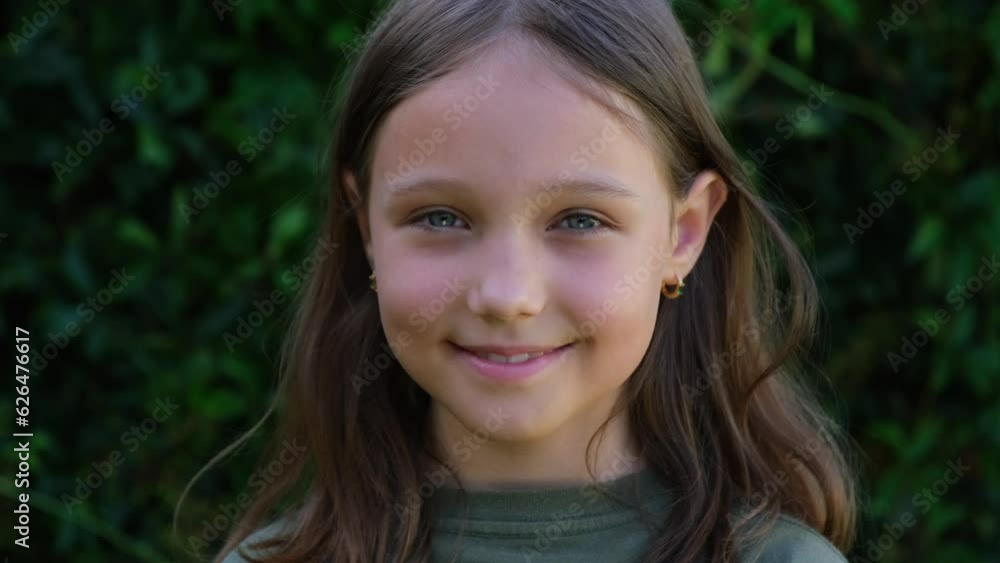 Portrait of a beautiful brunette little teenager girl looking at the camera and smiling, standing on the street against the backdrop of greenery on a summer day. Young curious child raises his eyes Portrait of a beautiful brunette little teenager girl looking at the camera and smiling, standing on the street against the backdrop of greenery on a summer day. Young curious child raises his eyes