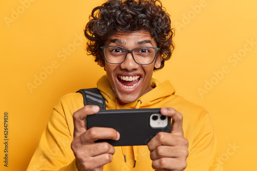 Wallpaper Mural People and positive emotions. Studio shot of young happy smiling Hindu male student standing in centre isolated on yellow background wearing casual hoodie excited to play favourite game on smartphone Torontodigital.ca