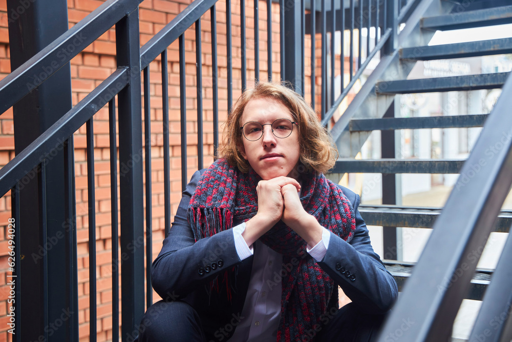 Fototapeta premium A handsome young man in a pantsuit and scarf. European. A college or university student. Street photo set. Sitting on the stairs