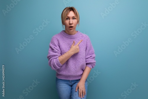 portrait of a shocked surprised blond young woman in a casual look shows her hand to the side on a bright background with copy space