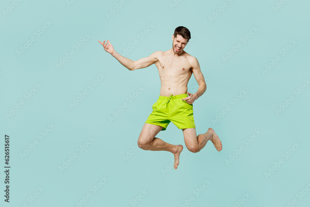 Full body young sexy man wear green shorts swimsuit relax near hotel pool jump high pretend playing guitar do arms gesture isolated on plain blue background. Summer vacation sea rest sun tan concept.