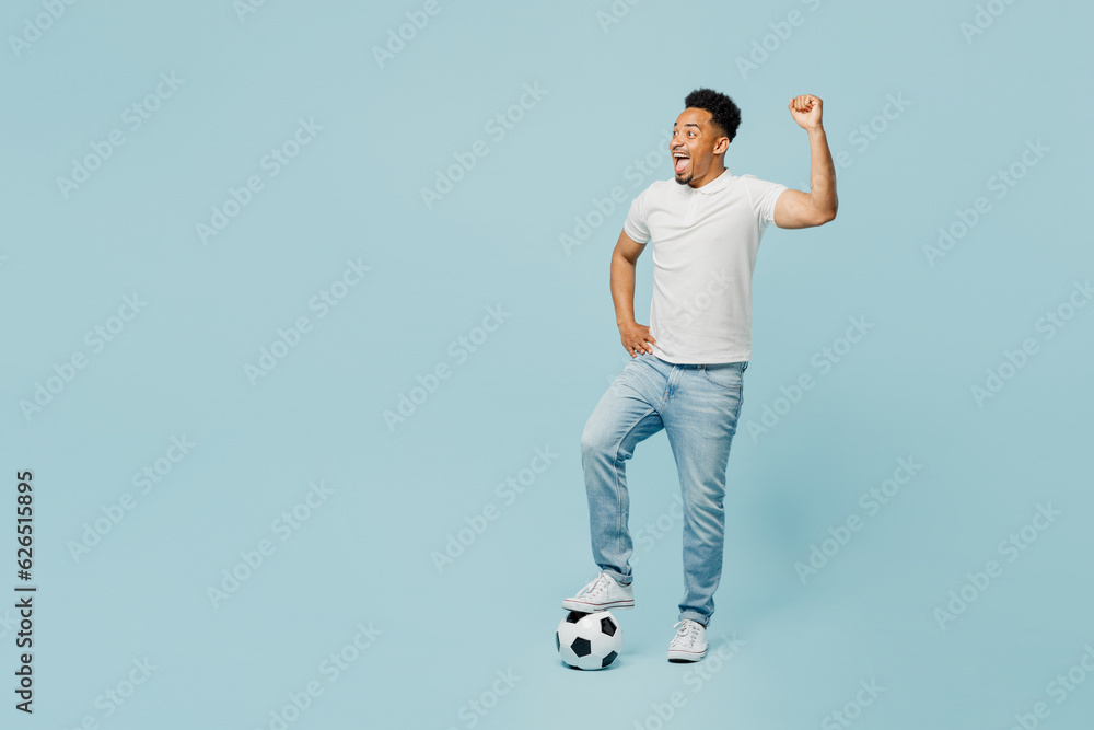 Full body sideways happy young man fan wear t-shirt cheer up support football sport team put leg on soccer ball watch tv live stream do winner gesture isolated on plain pastel blue color background. Full body sideways happy young man fan wear t-shirt cheer up support football sport team put leg on soccer ball watch tv live stream do winner gesture isolated on plain pastel blue color background.