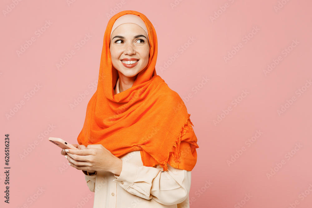 Side view young arabian asian muslim woman wear orange abaya hijab hold in hand use mobile cell phone look aside isolated on plain pink background. People uae middle eastern islam religious concept. Side view young arabian asian muslim woman wear orange abaya hijab hold in hand use mobile cell phone look aside isolated on plain pink background. People uae middle eastern islam religious concept.