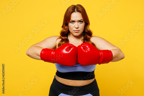 Young serious strict chubby overweight plus size big fat fit woman wear blue top red gloves warm up train boxing look camera isolated on plain yellow background studio home gym. Workout sport concept.
