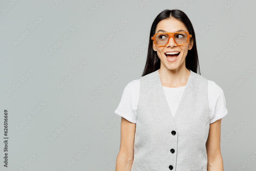 Young surprised shocked impressed astonished happy latin woman she wear white t-shirt gray vest glasses look aside on area mock up isolated on plain grey background studio portrait. Lifestyle concept. Young surprised shocked impressed astonished happy latin woman she wear white t-shirt gray vest glasses look aside on area mock up isolated on plain grey background studio portrait. Lifestyle concept.