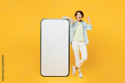 Full body young excited woman she wearing green t-shirt denim shirt casual clothes big huge blank screen mobile cell phone smartphone with area do winner gesture isolated on plain yellow background.