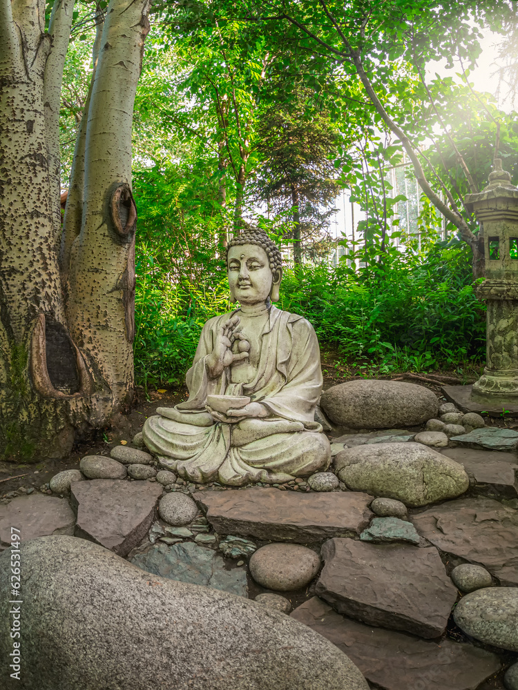 Stone Gautama Buddha decorative statue in a garden under green leaves. Outdoor Garden decor concept. Abakan, vertical view. Stone Gautama Buddha decorative statue in a garden under green leaves. Outdoor Garden decor concept. Abakan, vertical view.