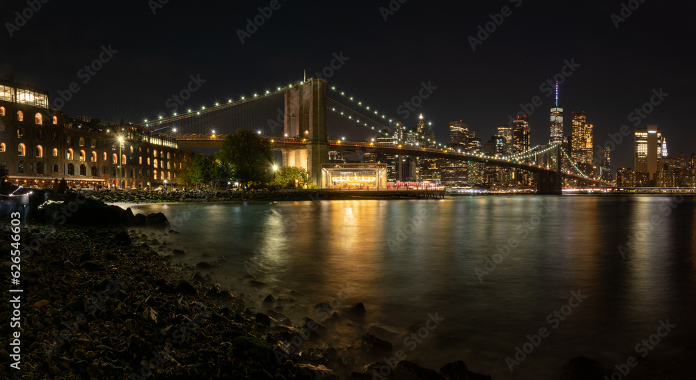 Brooklyn Bridge frames the Manhattan skyline at night, looking south from the dumbo section showing the full bridge with flags flying.