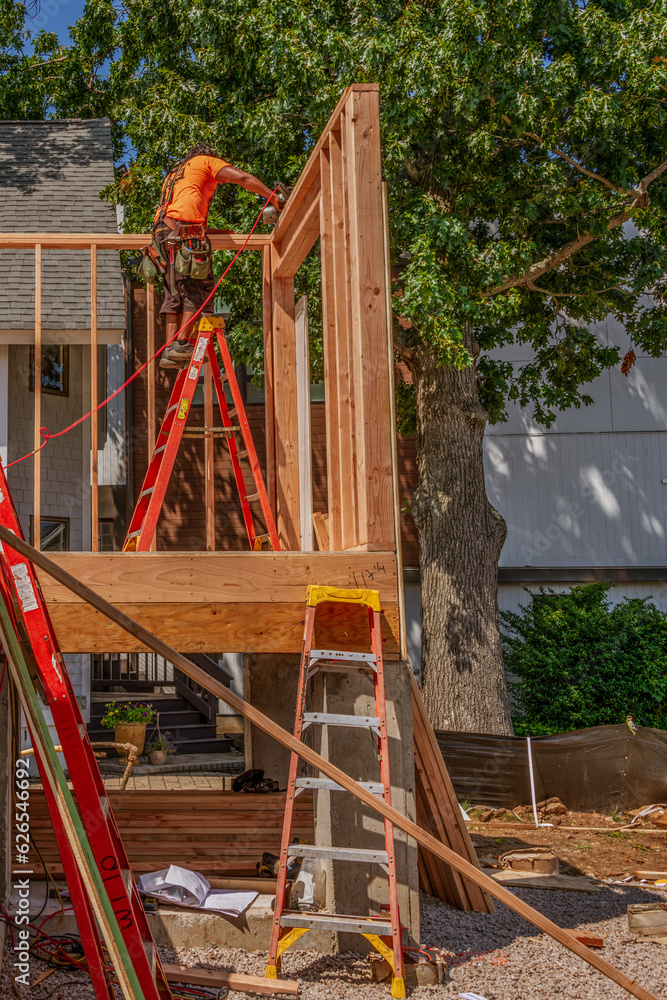 Framing the double top plate on the first floor of a home. The contractor standing on an aluminum ladder to reach the right position to use the nail gun to fasten the pieces Framing the double top plate on the first floor of a home. The contractor standing on an aluminum ladder to reach the right position to use the nail gun to fasten the pieces