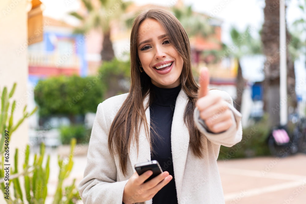 Young pretty woman at outdoors using mobile phone while doing thumbs up