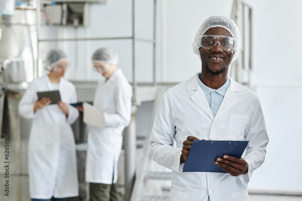 Waist up portrait of black young man wearing lab coat and smiling at camera in clean workshop of pharmaceutical factory, copy space