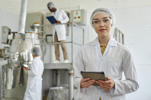 Waist up portrait of modern young woman wearing lab coat and looking at camera in clean workshop of pharmaceutical factory, copy space photo