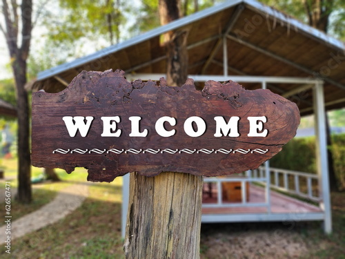 A large brown wooden sign written in white "WELCOME". A brown wooden sign is embroidered on a wooden pole above a large tree near the reception room. Signs are often installed in front of restaurants,