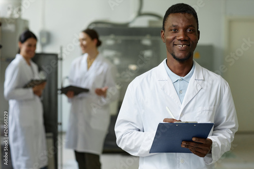 Waist up portrait of smiling black man wearing lab coat looking at camera in factory workshop and holding clipboard, copy space