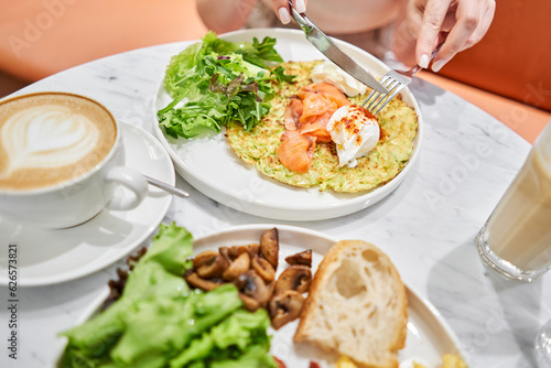 Woman cuts Zucchini pancakes, with avocado, cheese cream, salmon and egg . Healthy breakfast, protein. Restaurant dish. Breakfast in cafe on a sunny morning, on the summer veranda
