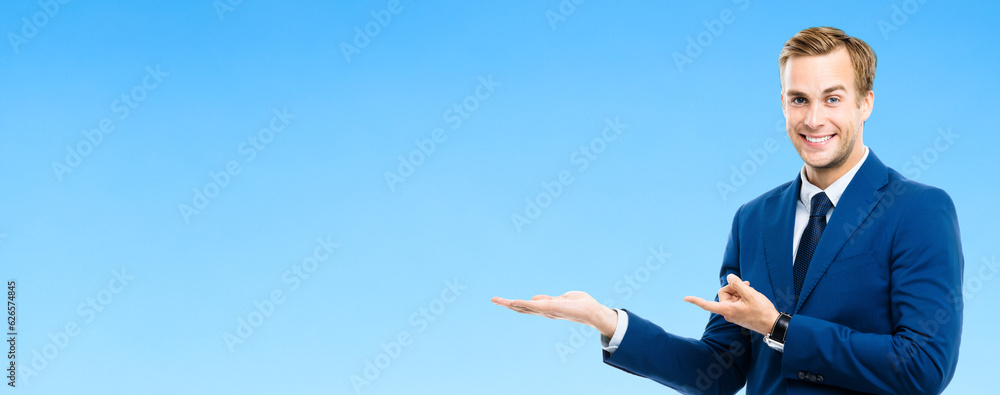 Portrait of confident businessman showing, giving or holding something, isolated on blue color background. Business success concept. Young happy smiling man at studio. Portrait of confident businessman showing, giving or holding something, isolated on blue color background. Business success concept. Young happy smiling man at studio.