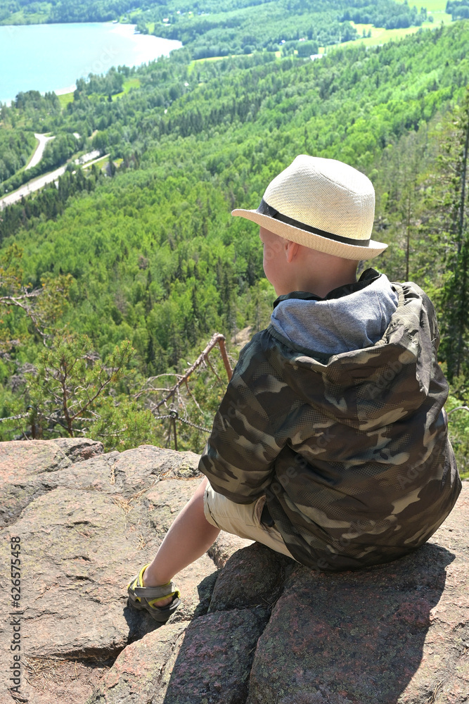 A boy in a hat sits relax high on a rock.