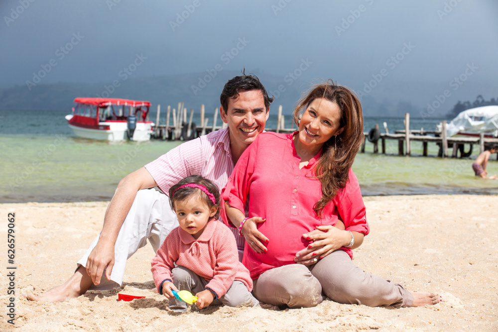 Fototapeta premium Young couple waiting for their second baby having fun with their baby girl at the beautiful white beach of Lake Tota located in the department of Boyaca at 3,015 meters above sea level in Colombia