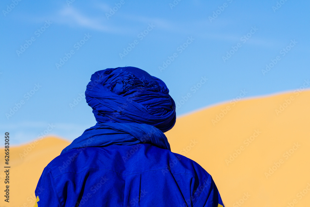 Close-up. Background. Berber in blue traditional clothing against the backdrop of a large dune. View from behind. Tunisia, Africa - obrazy, fototapety, plakaty 