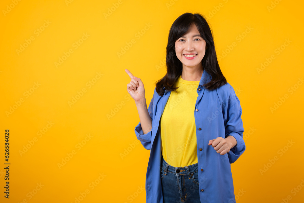 Capture attention with engaging portrait. A young Asian woman wearing a yellow t-shirt and blue shirt showing happy smile while pointing finger to free copy space. education promotion concept. Capture attention with engaging portrait. A young Asian woman wearing a yellow t-shirt and blue shirt showing happy smile while pointing finger to free copy space. education promotion concept.