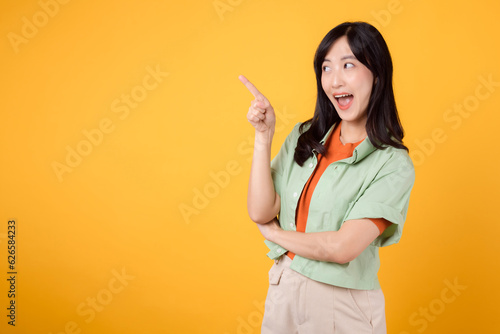 young Asian woman in her 30s, wearing a green shirt on an orange shirt. happy face and pointing finger to free copy space against vibrant yellow backdrop. Unlock savings and shop now!
