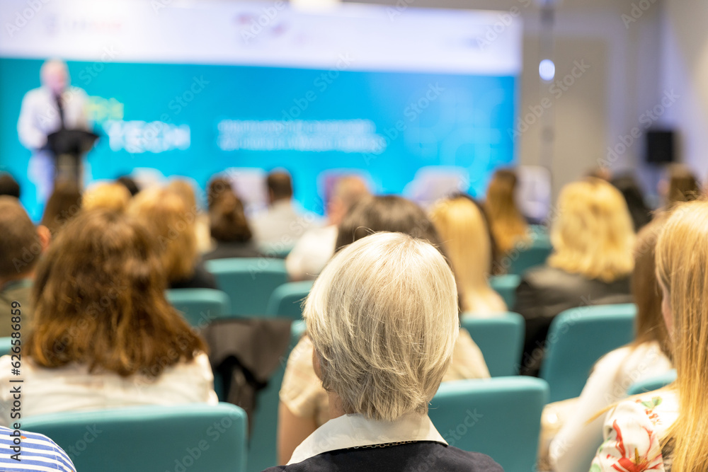 Fototapeta premium Rear view of participants at business conference, corporate presentation, workshop or life coaching training. Unrecognizable grey-haired woman in focus, blurred public speaker in the background.
