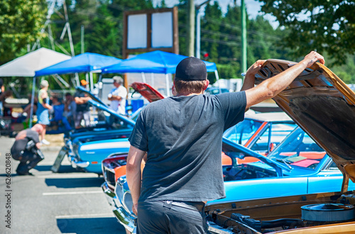 Man looks under the hood of vintage muscle car at classic automotive show.