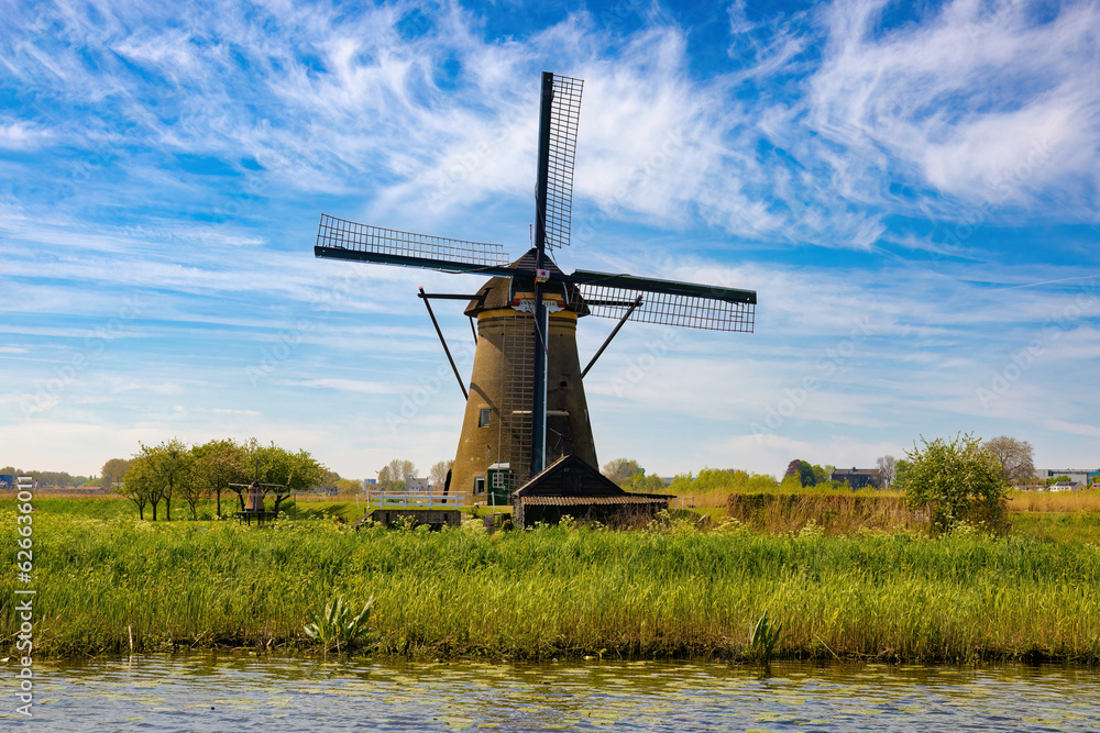 A large windmill on the other side of the channel stands like a great giant in the middle of the vast plain. Kinderdijk, The Netherlands.