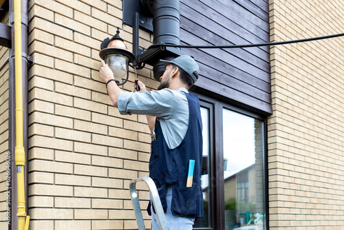 Young handsome electrician standing on ladder and change the light bulb in house facade on the backyard	