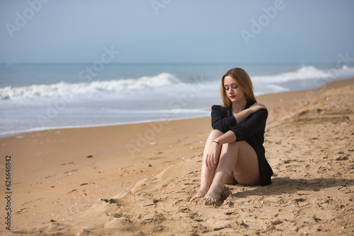 Young  blonde  beautiful woman in a bikini and black colored shirt sitting on the beach  sad  lonely  sorry  depressed. Concept loneliness  sadness  grief  depression.