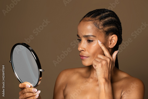 Skin aging. Attractive black middle aged woman holding mirror and looking at her wrinkles near eyes noticing age changes