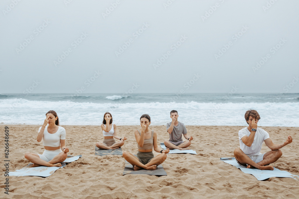 Group of women and men practising yoga on the beach, touching their foreheads with fingers, sitting on mats on sand