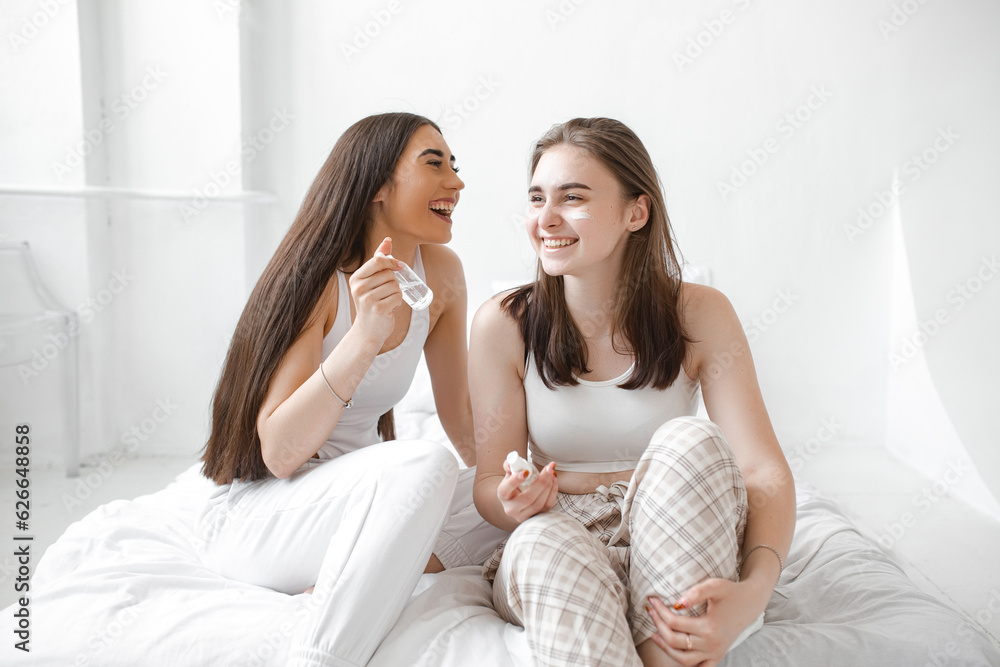 Two young beautiful girls with cosmetics sit on a white bed. Women use serum, cream and facial lotion to take care of their skin. Girlfriends in home spa rest and relax