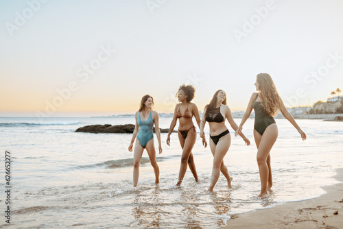 Happy young multiracial female friends in swimwear talking and laughing cheerfully while walking on the beach at sunset
